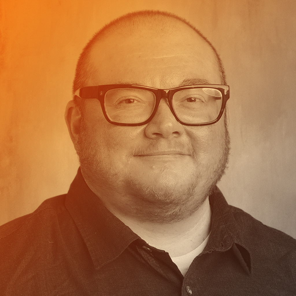 Ben's portrait. Ben is wearing a dark coloured shirt and glasses. He is smiling at the camera.