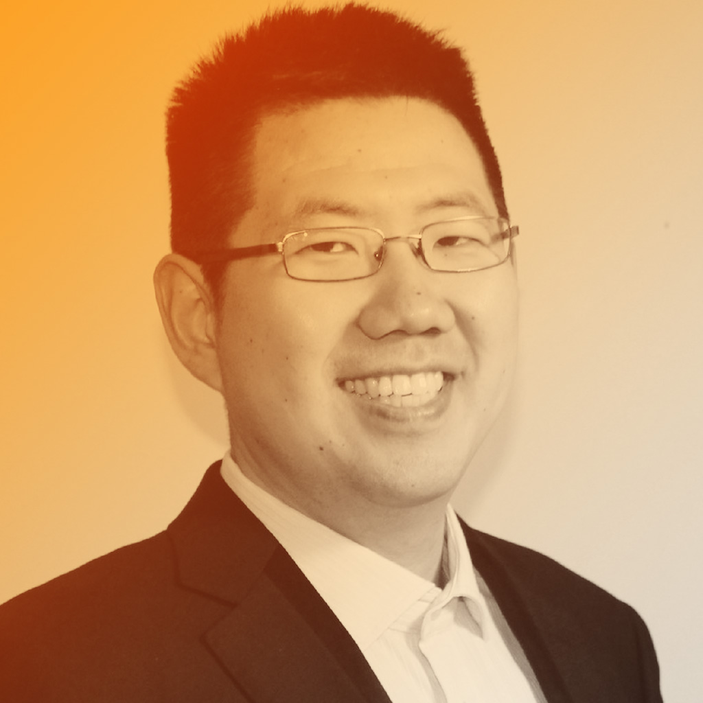 An asian man wearing glasses and black blazer, smiling at the camera.