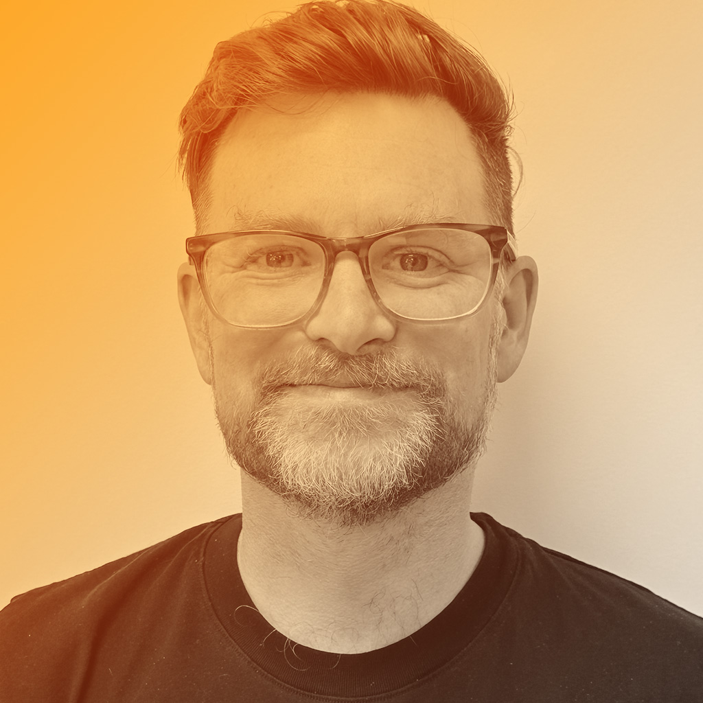 A middle aged man with a beard, glasses and a quiff hairstyle.