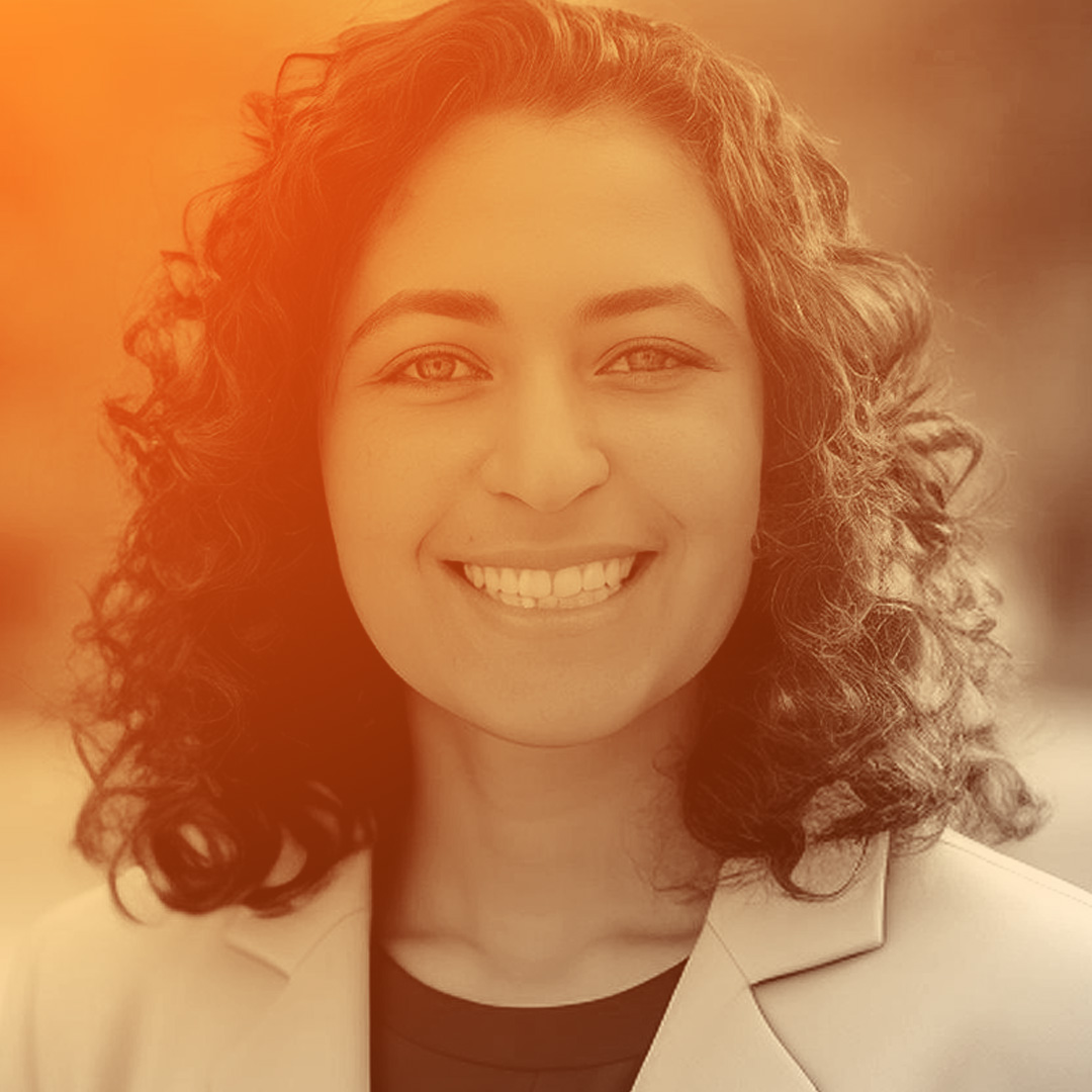Maia has brown curly hair, green eyes and is smiling broadly at the camera.