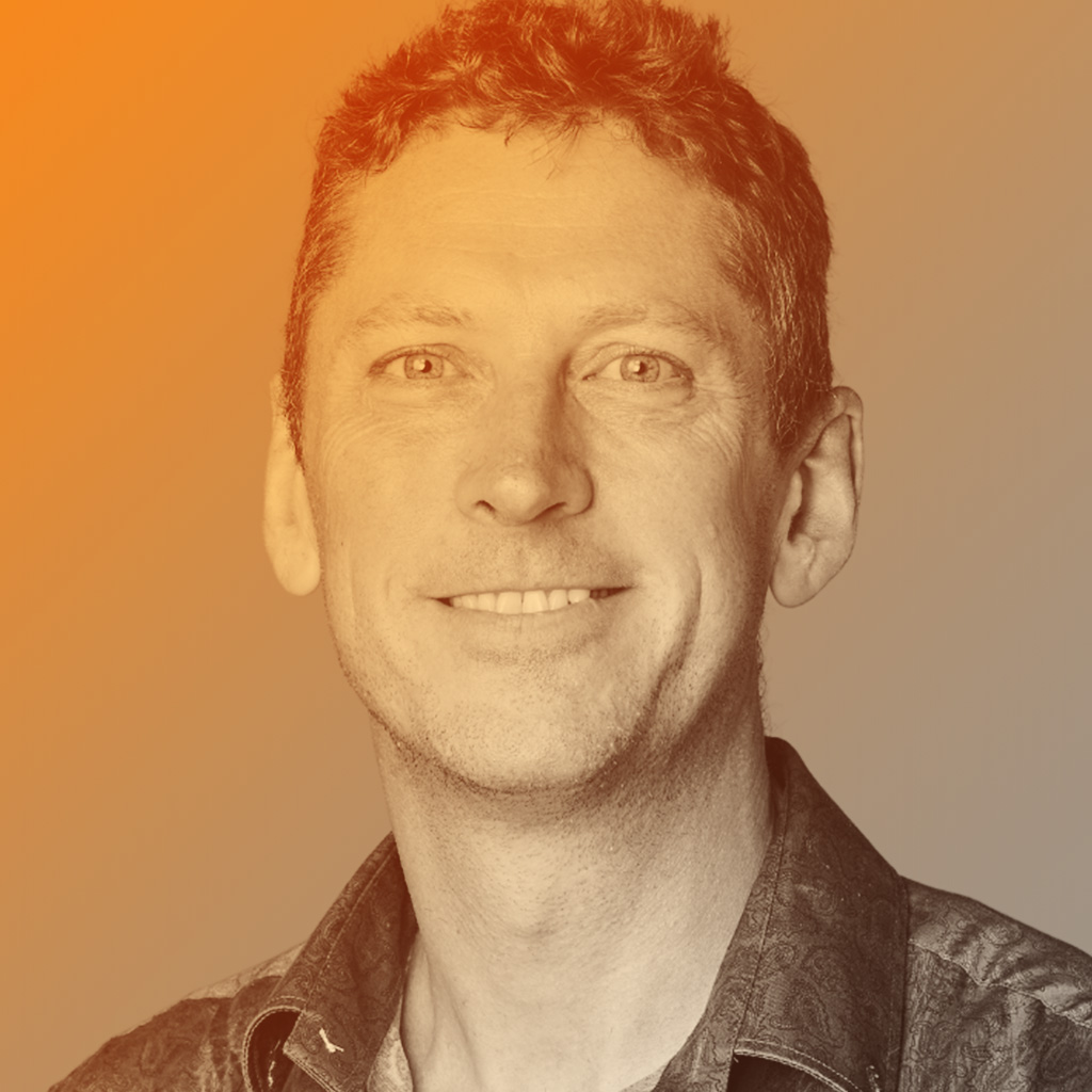 Smiling white man with short brown hair, wearing a patterned shirt, in front of a purple-to-green gradient background.