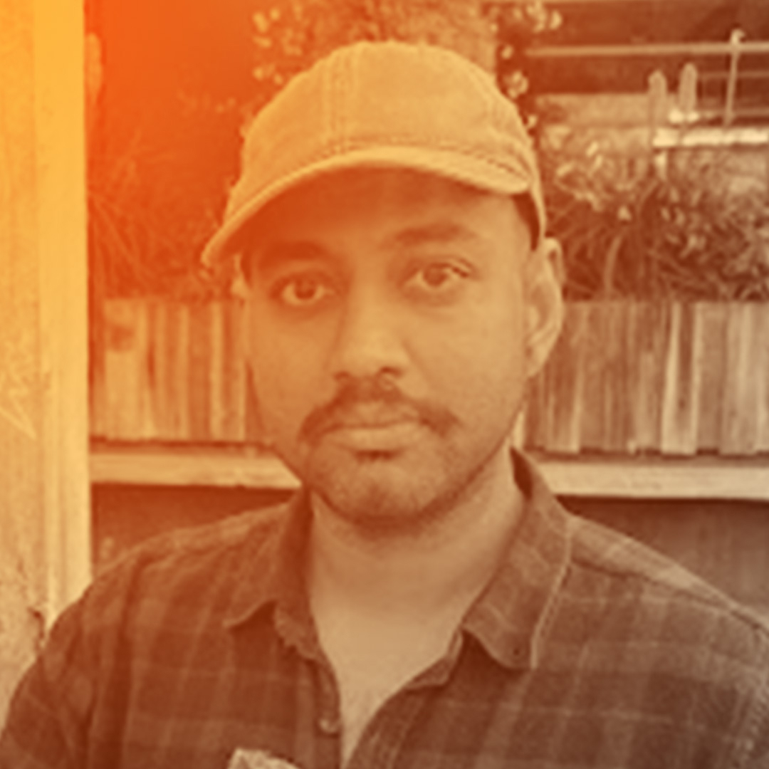 A mid 30’s Indian smiling at the camera while holding a tray of meatballs, one of which is delicately balanced on the end of a stick, now separated from its fellow meatball brethren. His outfit includes a blue tinged hat and dark blueish green flannel.