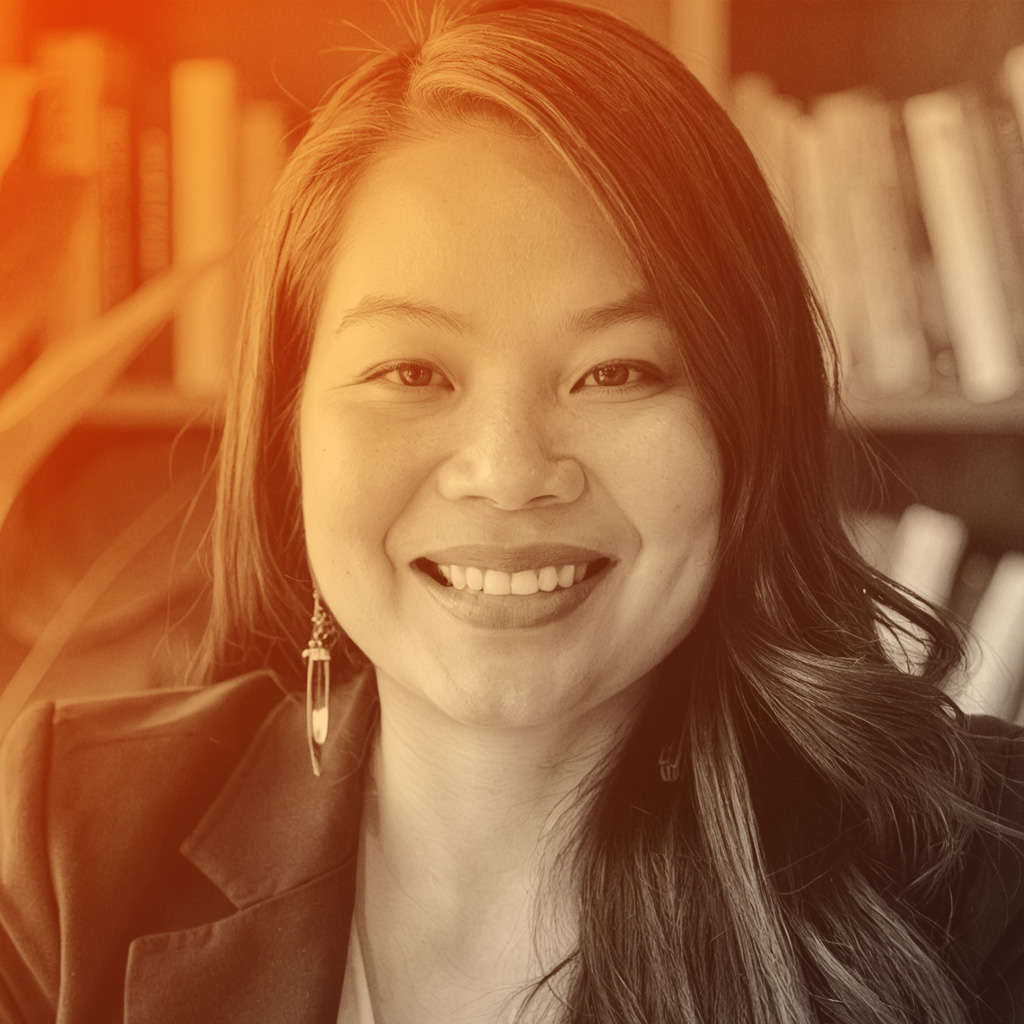 A woman with long dark hair smiles warmly at a desk. She wears a black blazer over a white top, with books and plants in the background. 
